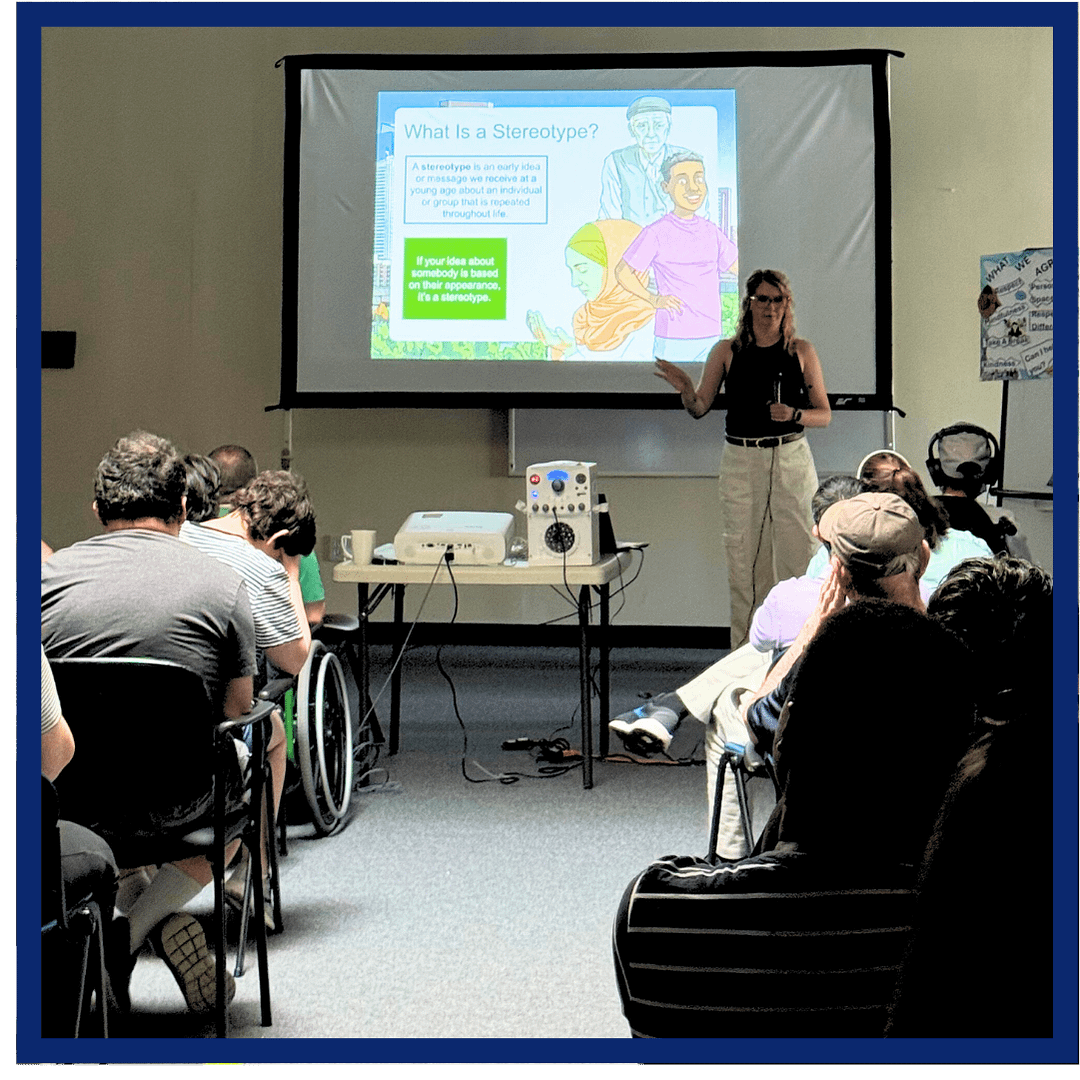 Melissa (in a black tank top and white pants) stands in front of a room of students, teaching with a powerpoint presentation behind her.