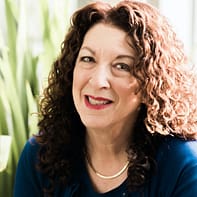 Shelly sits in front of a green background, wearing a blue shirt and gold necklace. She is smiling and has curly dark brown hair.