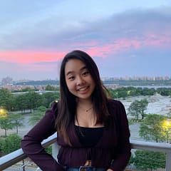 Jennifer Lee stands on a balcony with a sunset over a leafy cityscape by river in the background. She is smiling at the camera, has long brown hair, and is wearing a purple top.