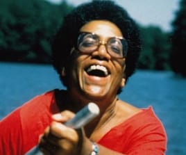 Audre Lorde smiles joyously toward the camera. She is wearing large wide rimmed glasses, and a red top. She is in a natural setting with a background of water and forest.