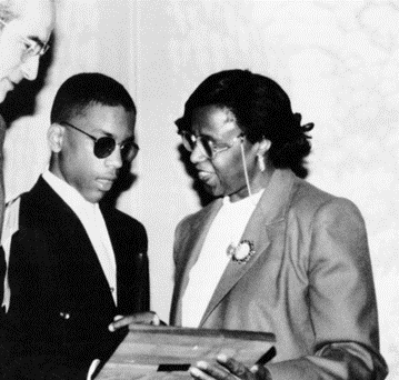 A black and white photograph of Sylvia Walker, a black woman wearing a blazer with a pin on the lapel and glasses, receiving an award and shaking hands with a teenaged African American student who is blind and wearing a black blazer and white shirt and tie.
