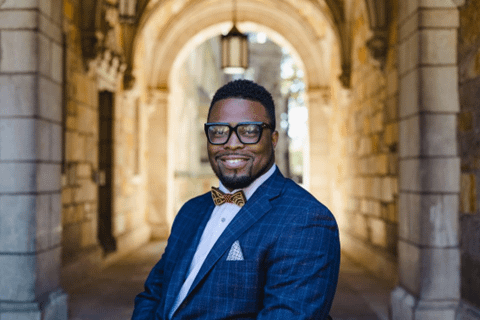 Dr. Oluwaferanmi Okanlami is photographed wearing a dark blue check suit with light blue shirt and matching pocket square. He is wearing an elegant red and gold bowtie, has a goatee and large black frame glasses. The background is an elegant stone building entrance with a decorative lantern.