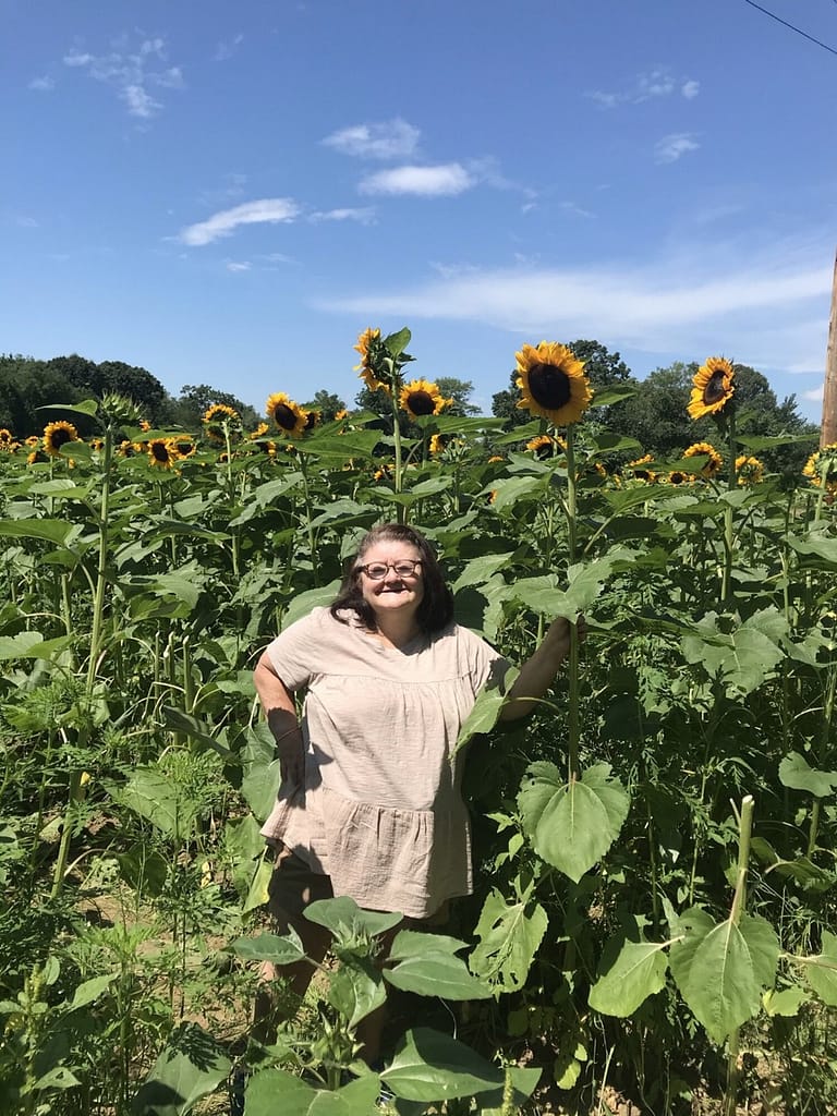 Donna Jay, a white woman with shoulder length brown hair, wears glasses and a tan dress standing in a field of sunflowers smiling at the camera.