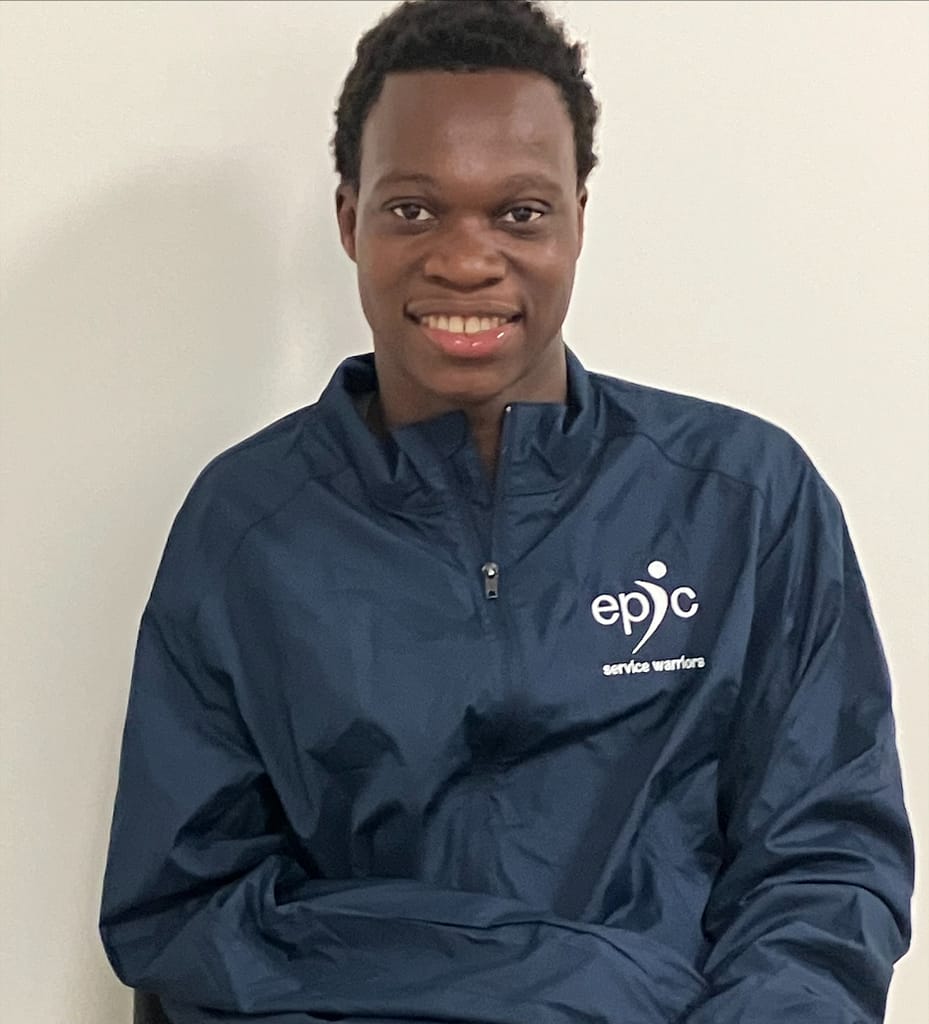 Ewidji sits in front of a white wall. He is smiling, has short black hair, and is wearing a navy blue EPIC jacket.